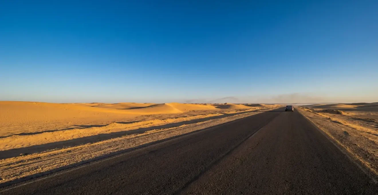 Route goudronnée traversant un paysage désertique avec dunes en arrière-plan